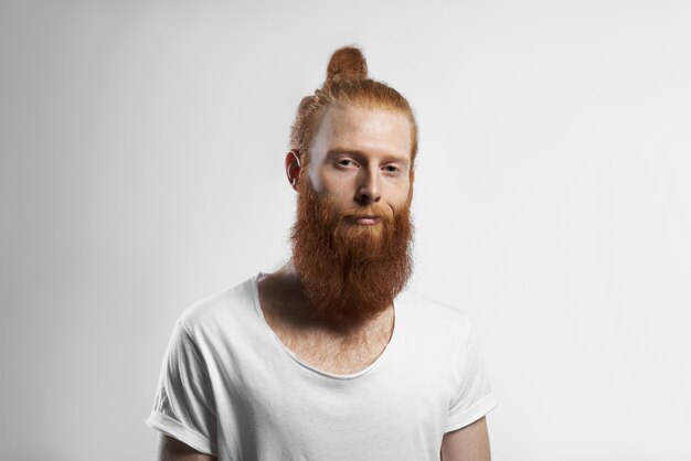 Menschen, Jugend, Stil und Modekonzept. Bild des schönen positiven trendy aussehenden jungen rothaarigen Hipster-Mannes mit unscharfem Bart und Haarknoten, der im Studio aufwirft, gekleidet in lässigem weißem T-Shirt