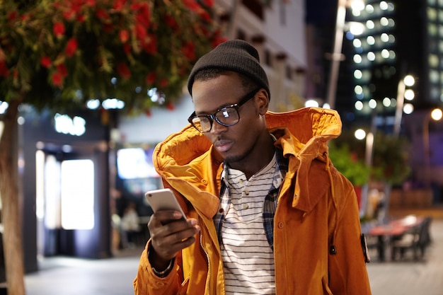 Menschen, Lebensstil, Reisen, Tourismus und moderne Technologie. Müder junger Afroamerikaner, der Handy verwendet, um Taxi über Online-Taxiservice-App anzufordern, um nach langem Flug zum Hotel zu gelangen