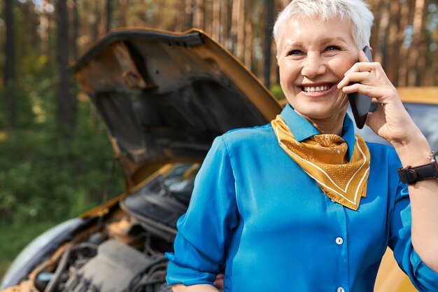 Menschen, Lebensstil, Transport und modernes Technologiekonzept. Schöne blonde pensionierte Frau, die durch gebrochenes Auto mit offener Motorhaube steht, Pannenhilfe ruft, um Hilfe bittet, lächelt