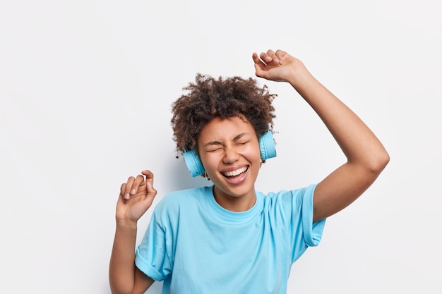 Menschen Lifestyle-Freizeitkonzept. Energiegeladene, fröhliche Afroamerikanerin mit lockigem Haar tanzt unbeschwert hebt die Arme, fühlt sich amüsiert genießt Lieblingsmusik trägt drahtlose Kopfhörer blaues T-Shirt