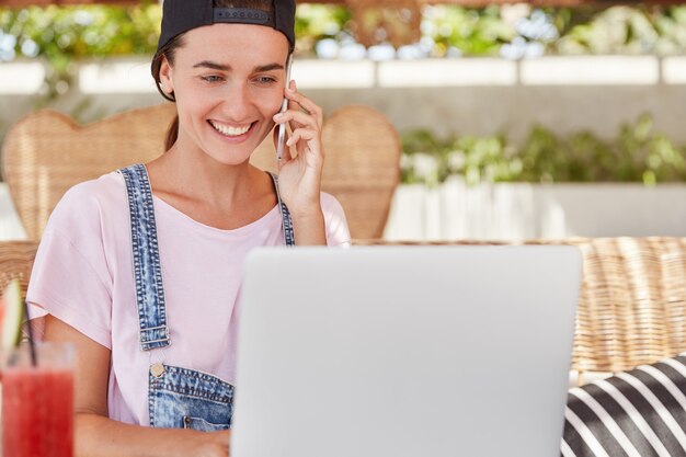 Menschen-, Lifestyle- und Technologiekonzept. Schöne lächelnde Frau in stilvoller Mütze und Jeansoverall teilt gute Nachrichten über Smartphone, Tastaturinformationen auf Laptop-Computer, sitzt im Café