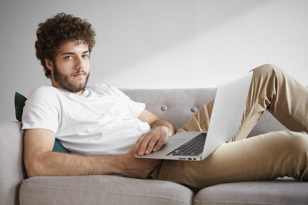 Menschen, moderne Technologie und Kommunikationskonzept. Bild des hübschen stilvollen Kerls mit Bart, der auf Sofa mit tragbarem Computer auf seinem Schoß sitzt und Hochgeschwindigkeits-drahtlose Internetverbindung genießt