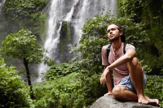 Menschen, Natur und Abenteuer. Junger Hipster mit Rucksack, der auf großem Felsen durch Wasserfall sitzt