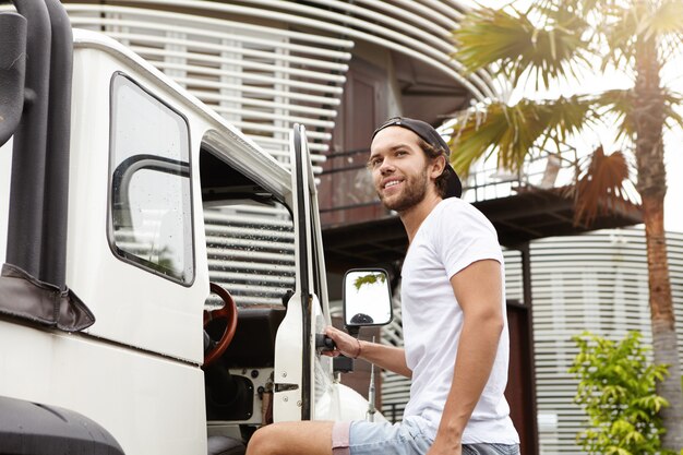 Menschen-, Reise- und Abenteuerkonzept. Attraktiver Mann mit Bart, der nach einem aktiven Wochenende im Freien lächelt und sich von seinen Freunden verabschiedet, während er vor seinem weißen Jeep steht