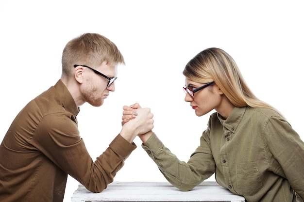 Kostenloses Foto menschen, teamwork, kooperation und wettbewerbskonzept. seitenansicht von jungen weiblichen und bärtigen männlichen kollegen, die beide eine brille tragen und sich mit selbstbewussten, entschlossenen blicken anstarren