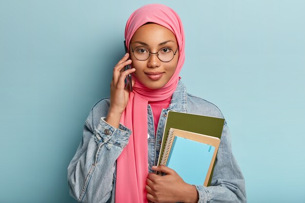 Menschen, Technologie, ethnische Zugehörigkeit, Kommunikationskonzept. Hübsches Mädchen im traditionellen muslimischen Hijab hat Telefongespräch mit Gruppenmitglied, bespricht zukünftiges Projekt, hält zwei Spiralblöcke, posiert drinnen