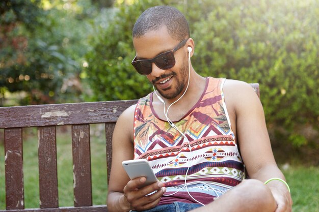 Menschen, Technologie, Freizeit und Lifestyle - glückliche Hipster-Studenten, die mit dem Smartphone im Internet surfen und sich im Freien entspannen. Junger Freiberufler, der Kopfhörer trägt, sitzt auf Bank, die allein entspannt