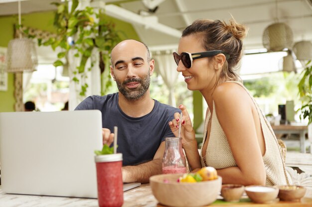 Menschen, Technologie und Kommunikation. Erwachsenes Paar mit Laptop-Computer im Café, am Tisch mit frischen Getränken sitzend. Hübscher Mann, der seiner Freundin etwas auf Notizbuch zeigt.