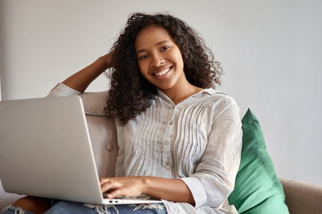 Menschen, Technologie und Kommunikation. Hübsches Mädchen mit voluminöser Afro-Frisur, die auf Couch mit tragbarem elektronischem Gerät auf ihrem Schoß sitzt, online einkauft oder Lieblingsserie beobachtet, lächelt