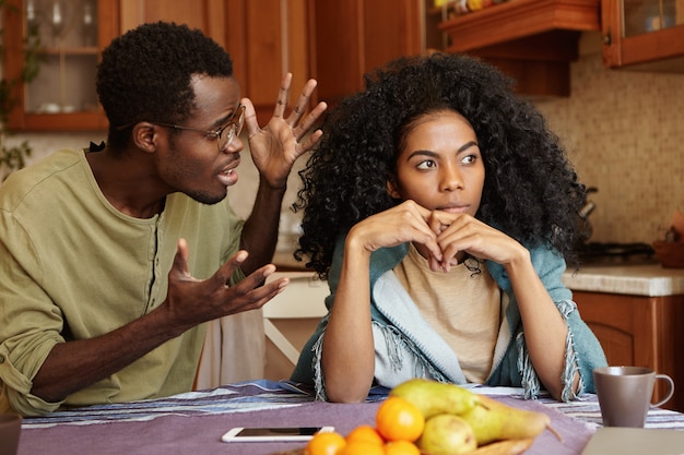 Menschen- und Beziehungskonzept. Afroamerikanisches Paar, das in der Küche argumentiert: Mann in Gläsern, der vor Wut und Verzweiflung gestikuliert und seine schöne unglückliche Freundin anschreit, die ihn völlig ignoriert