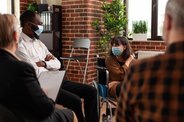 Menschen und Frauen sitzen im Rollstuhl und unterhalten sich bei einem Treffen. Multiethnische Patienten, die während der Covid-19-Pandemie ein Gespräch mit einem Psychiater bei einer Selbsthilfegruppentherapie besuchen.