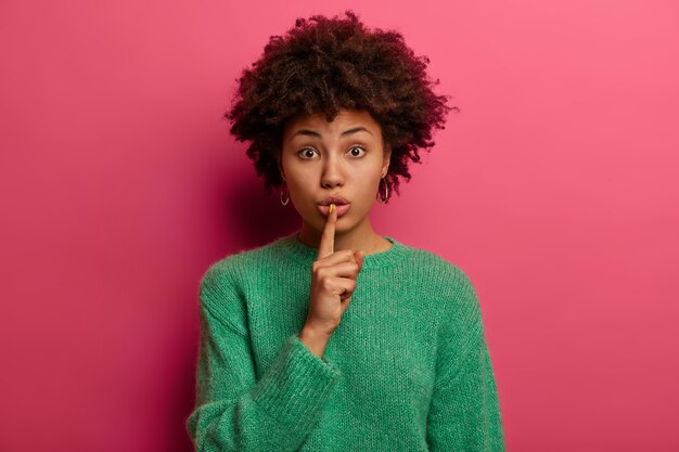 Menschen und Geheimhaltung. Teenager drückt Zeigefinger an die Lippen, erzählt vertrauliche Informationen, klatscht über etwas, steht stumm, sieht geheimnisvoll aus, trägt einen grünen Pullover, steht drinnen