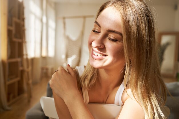 Menschen- und Lifestyle-Konzept. Innenaufnahme des freudigen hübschen jungen Mädchens mit dem glatten blonden Haar, das die Rückenlehne des Stuhls umarmt, die rückwärts sitzt und die Augen wegen des hellen Sonnenscheins schließt und breit lächelt