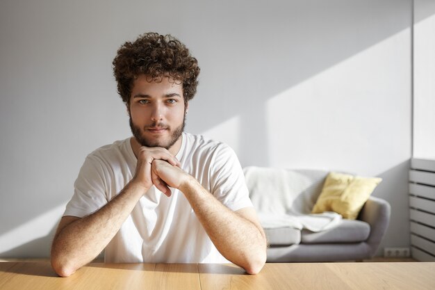 Menschen- und Lifestyle-Konzept. Innenaufnahme des positiven gutaussehenden jungen kaukasischen Mannes mit der voluminösen Frisur und dem dicken Bart lächelnd, Freizeit zu Hause verbringend, lokal am Holztisch sitzend