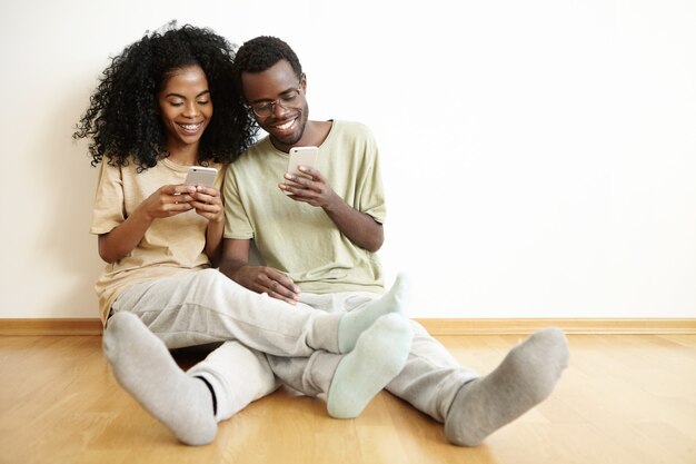 Menschen und moderne Technik. Lässiges, gemütliches afrikanisches Paar, das Zeit miteinander verbringt, online einzukaufen oder Apps für Gadgets zu verwenden, kostenloses WLAN zu Hause zu genießen und auf dem Boden zu sitzen