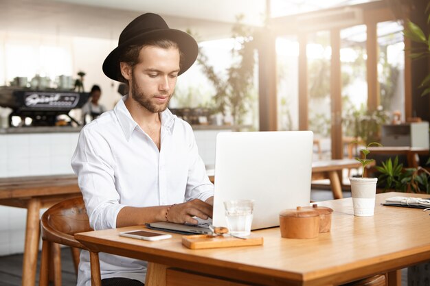 Menschen, Unternehmen und moderne Technologie. Ernsthafter und konzentrierter gutaussehender Mann, der während des Frühstücks am Kaffeetisch mit Glas Wasser und Handy sitzt und Hände auf der Tastatur seines generischen Laptops hält