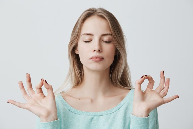 Menschen, Yoga und gesunder Lebensstil. Wunderschöne junge blonde Frau in hellblauem Pullover, der die Augen geschlossen hält, während er drinnen meditiert, Seelenfrieden übt und Finger in der Mudra-Geste hält