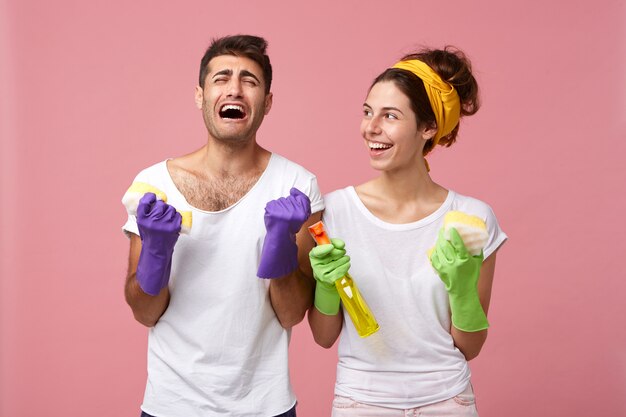 Menschliche Emotionen, Gefühle und Einstellungen. Unglücklicher bärtiger Mann in Gummihandschuhen, der weint und sich über Hausarbeit gestresst fühlt, während glückliche Frau neben ihm mit Putzzeug steht und lacht