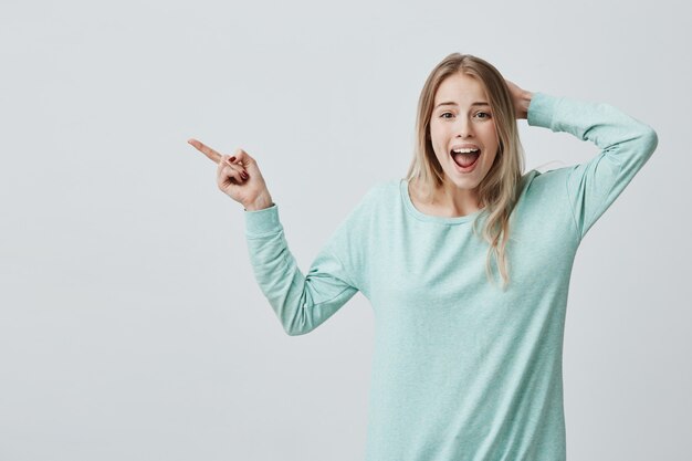 Menschliche Gesichtsausdrücke, Emotionen und Gefühle. Erstaunt schockierte junge blonde Frau in Freizeitkleidung, die mit dem Zeigefinger auf eine leere Wand zeigte, überrascht von den Verkaufspreisen und den Mund weit offen hielt