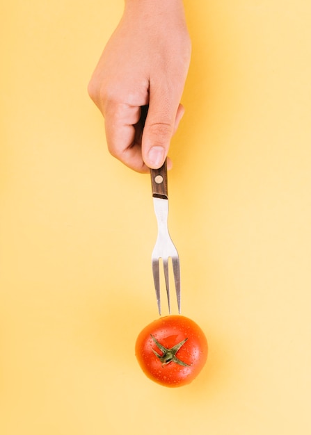 Menschliche Hand, die Gabel in rote Tomate auf gelbem Hintergrund einfügt