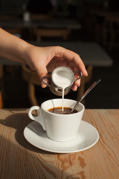 Menschliche Hand, die Milch in den schwarzen Kaffee in der Cafeteria gießt