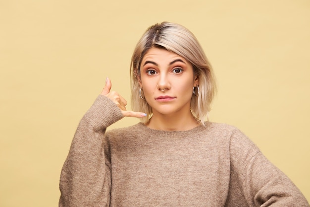 Menschliche Mimik und Körpersprache. Positive gut aussehende junge blonde Frau hält Hand an seinem Ohr imitiert Handy, sagt Rufen Sie mich an, ich werde in Kontakt sein, isoliert posieren