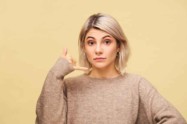 Menschliche Mimik und Körpersprache. Positive gut aussehende junge blonde Frau hält Hand an seinem Ohr imitiert Handy, sagt Rufen Sie mich an, ich werde in Kontakt sein, isoliert posieren