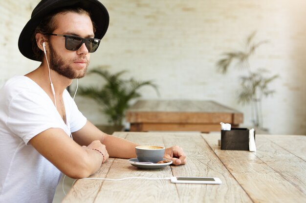 Menschliche und moderne Technologie. Modischer junger bärtiger Mann, der stilvollen Hut und schwarze Schatten trägt, die am Terrassencafé sich entspannen und am Tisch mit Becher sitzen