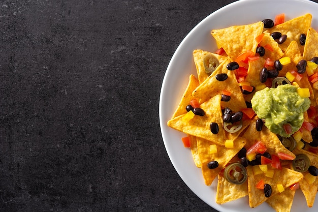 Mexikanische Nachos-Tortilla-Chips mit Guacamole-Tomaten und Jalapeno aus schwarzen Bohnen auf schwarzem Hintergrund