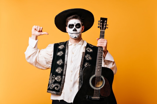 Kostenloses Foto mexikanischer mann mit hut zeigt mit dem finger auf die gitarre. schnappschuss des kerls im traditionellen kleid mit gesichtskunst auf isolierter wand.