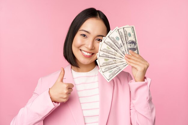 Mikrokreditinvestitionen und Geschäftsleute Konzept Junge asiatische Geschäftsfrau Corporate Lady zeigt Geld Bargeld Dollar Daumen hoch und empfiehlt Unternehmen rosa Hintergrund