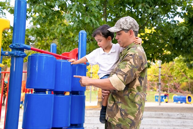 Militärvater, der mit kleinem Sohn im Park geht, Jungen in den Armen hält und Spielgeräte studiert. Seitenansicht. Elternschafts- oder Kindheitskonzept