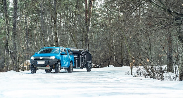 Mini-Wohnmobil auf der Winterwaldstraße, die von einem blauen Muskelauto gezogen wird Wohnwagen oder Wohnmobil-Wohnmobilanhänger auf einer Bergstraße Familienurlaub und Reisekonzept