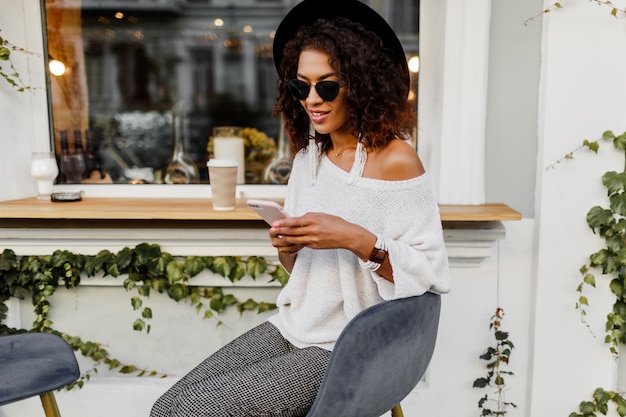 Mischen Sie Rennfrau im stilvollen lässigen Outfit, das im Stadtcafé im Freien entspannt, Kaffee trinkt und per Handy plaudert. Trägt trendige Accessoires und Sonnenbrillen.