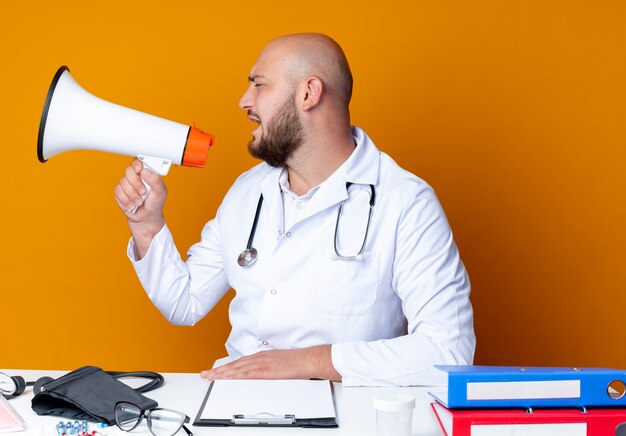 Mit Blick auf den jungen kahlen männlichen Arzt der Seite, der medizinische Robe und Stethoskop trägt, die am Schreibtisch mit medizinischen Werkzeugen sitzen, spricht auf Lautsprecher lokalisiert auf orangefarbenem Hintergrund