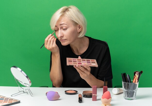 Mit Blick auf den Spiegel sitzt das junge schöne Mädchen am Tisch mit Make-up-Tools, die Lidschatten mit Make-up-Pinsel auf der grünen Wand auftragen