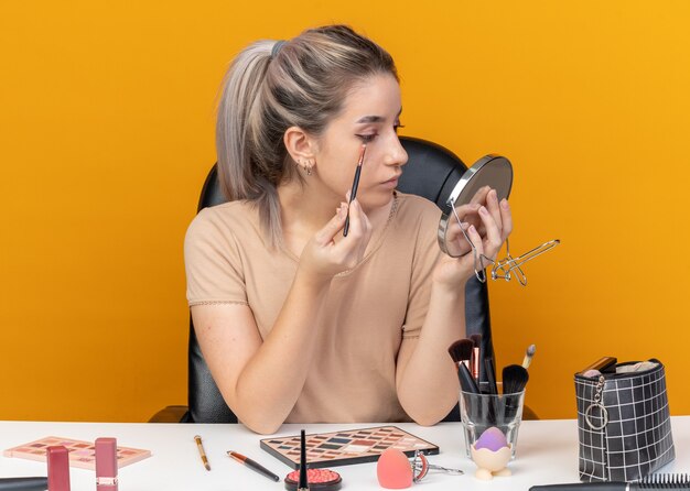 Mit Blick auf den Spiegel sitzt das junge schöne Mädchen am Tisch mit Make-up-Tools, die Lidschatten mit Pinsel auf der orangefarbenen Wand auftragen