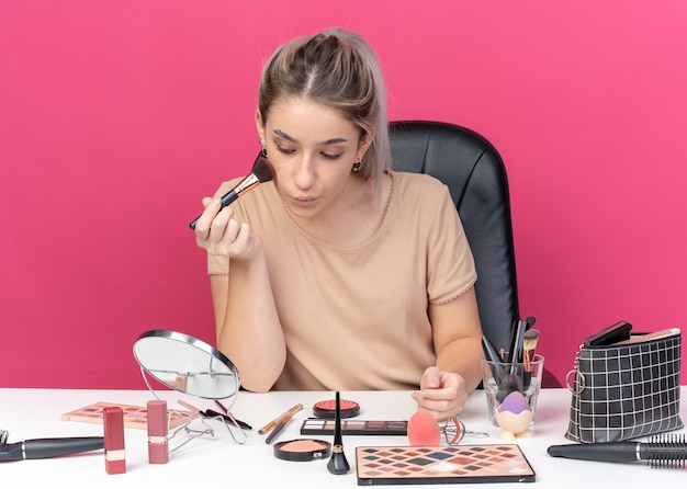 Mit Blick auf den Spiegel sitzt das junge schöne Mädchen am Tisch mit Make-up-Tools, die Puderrouge einzeln auf rosa Wand auftragen