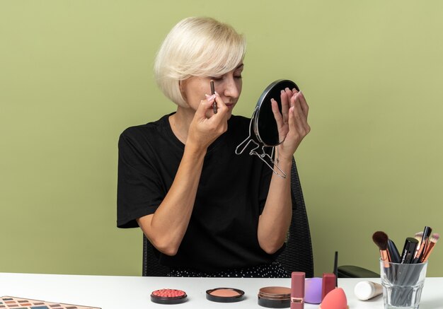 Mit Blick auf den Spiegel sitzt das junge schöne Mädchen am Tisch mit Make-up-Tools und zeichnet Pfeil mit Eyeliner isoliert auf olivgrüner Wand