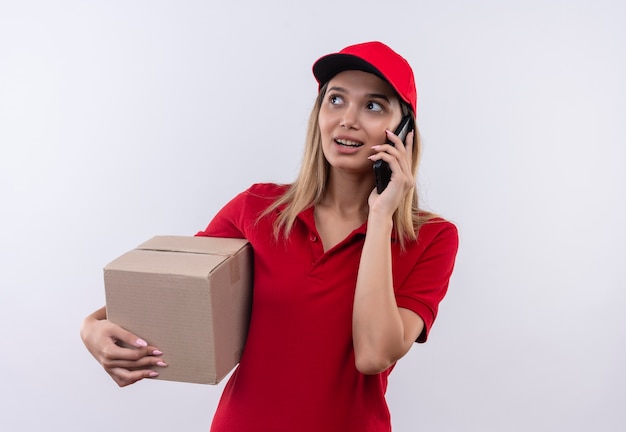 Mit Blick auf die Seite erfreut junges Liefermädchen, das rote Uniform und Kappenhaltebox trägt und am Telefon lokalisiert auf weißer Wand spricht