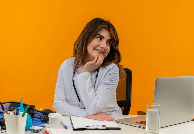 Mit Blick auf die Seite erfreut Ärztin mittleren Alters in medizinischer Robe mit Stethoskop, die am Schreibtisch sitzt, arbeiten am Laptop mit medizinischen Werkzeugen, die Hand auf Kinn auf orange Wand mit Kopienraum setzen