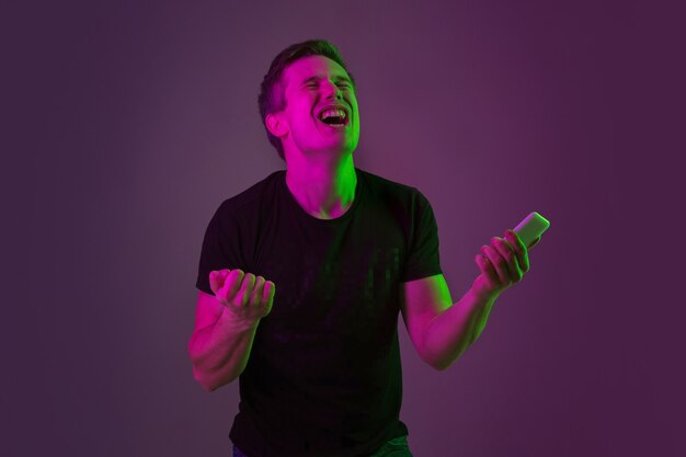 Mit dem Smartphone wetten, gewinnen. Porträt des kaukasischen Mannes auf purpurrotem Studiohintergrund im Neonlicht. Schönes männliches Modell im schwarzen Hemd. Konzept der menschlichen Emotionen, Gesichtsausdruck, Verkauf, Anzeige.