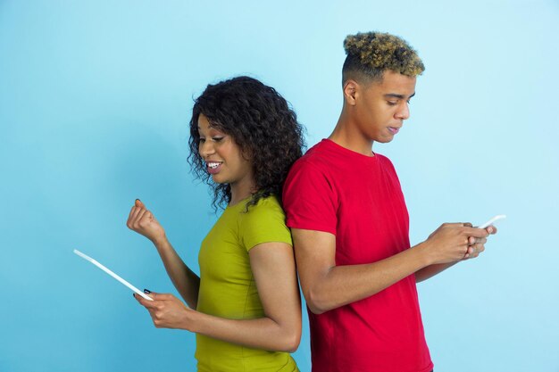 Mit Gadgets lachen. Junger emotionaler afroamerikanischer Mann und Frau in den bunten Kleidern auf blauem Hintergrund.