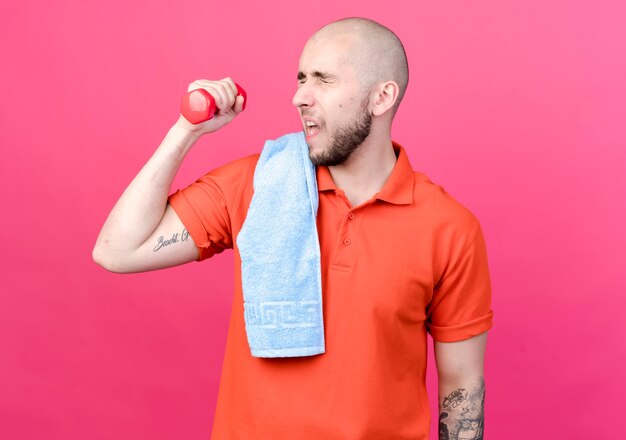 Mit geschlossenen Augen angespannter junger sportlicher Mann, der mit Hantel mit Handtuch auf Schulter auf rosa Wand isoliert trainiert