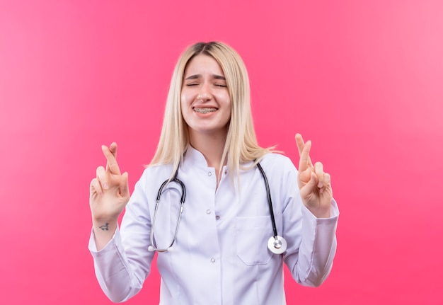 Mit geschlossenen Augen besorgt Doktor junges blondes Mädchen, das Stethoskop im medizinischen Kleid und in der Zahnspange trägt, die Finger auf isolierter rosa Wand kreuzen