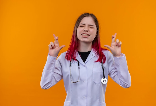 Mit geschlossenen Augen betroffenes junges Ärztinmädchen, das medizinisches Gewand des Stethoskops trägt, das Finger auf isolierter orange Wand kreuzt