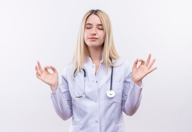 Mit geschlossenen Augen Doktor junges blondes Mädchen, das Stethoskop und medizinisches Kleid trägt, zeigt okey Geste mit beiden Händen auf isolierter weißer Wand
