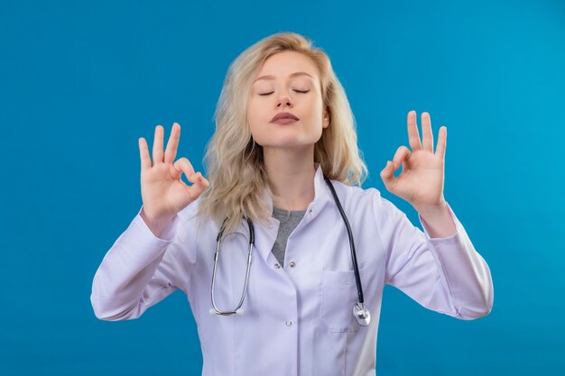 Mit geschlossenen Augen Doktor junges Mädchen, das Stethoskop im medizinischen Kleid trägt, das okey Geste auf blauem Hintergrund zeigt