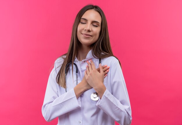 Mit geschlossenen Augen erfreut junge Ärztin Mädchen im Stethoskop medizinischen Kleid legte ihre Hände auf Herz auf isolierte rosa Wand