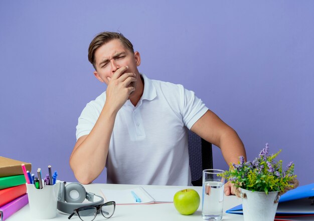 Mit geschlossenen Augen gähnt junger hübscher männlicher Student, der am Schreibtisch mit Schulwerkzeugen sitzt, bedeckte Mund mit Hand lokalisiert auf Blau